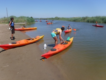 Kayakistas en la orilla de un río o canal, listos para iniciar o continuar su recorrido, con varias embarcaciones ya en el agua.