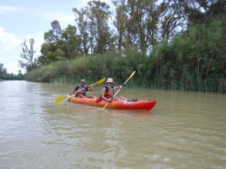 Dos personas disfrutan de un paseo en kayak biplaza por un río, rodeados de vegetación densa y árboles altos a lo largo de las orillas.