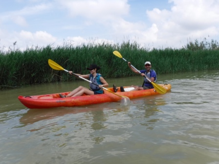 Dos personas disfrutan de un tranquilo paseo en kayak por un río, remando en tándem junto a una orilla con vegetación alta.