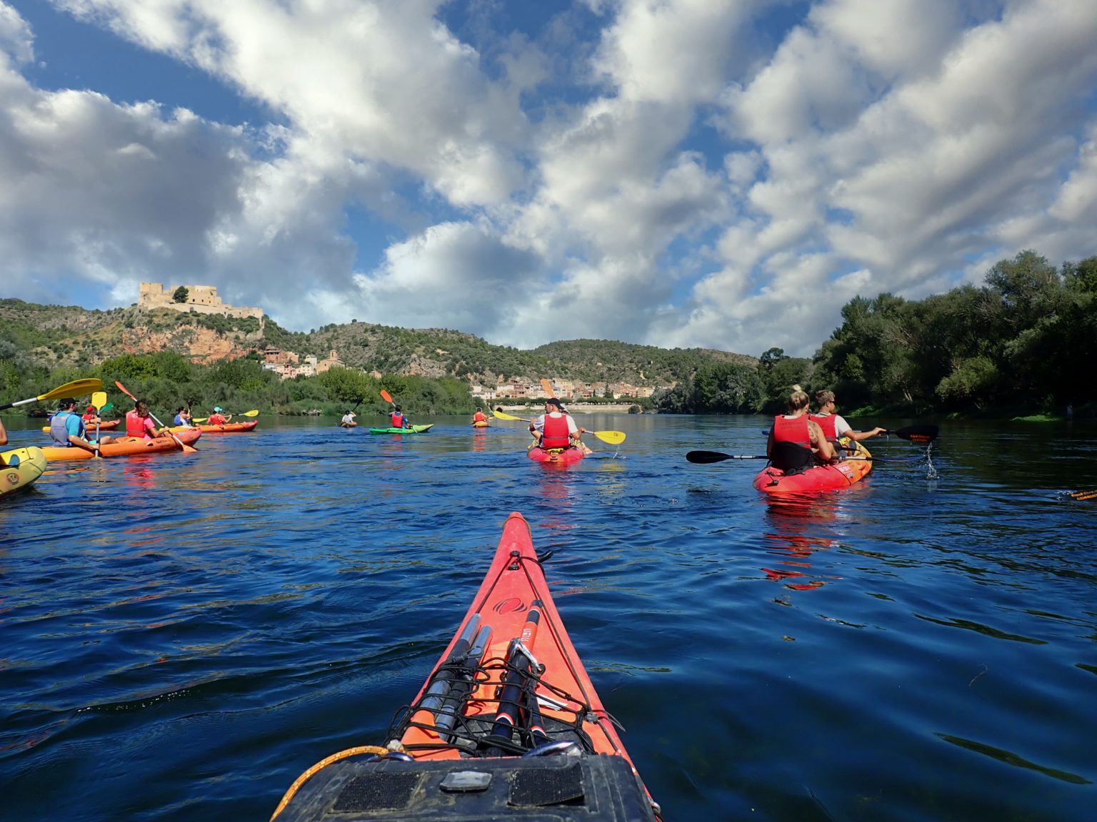 Un grupo de kayakistas disfruta de un tranquilo descenso por el río Ebro, con el majestuoso Castillo de Miravet al fondo.