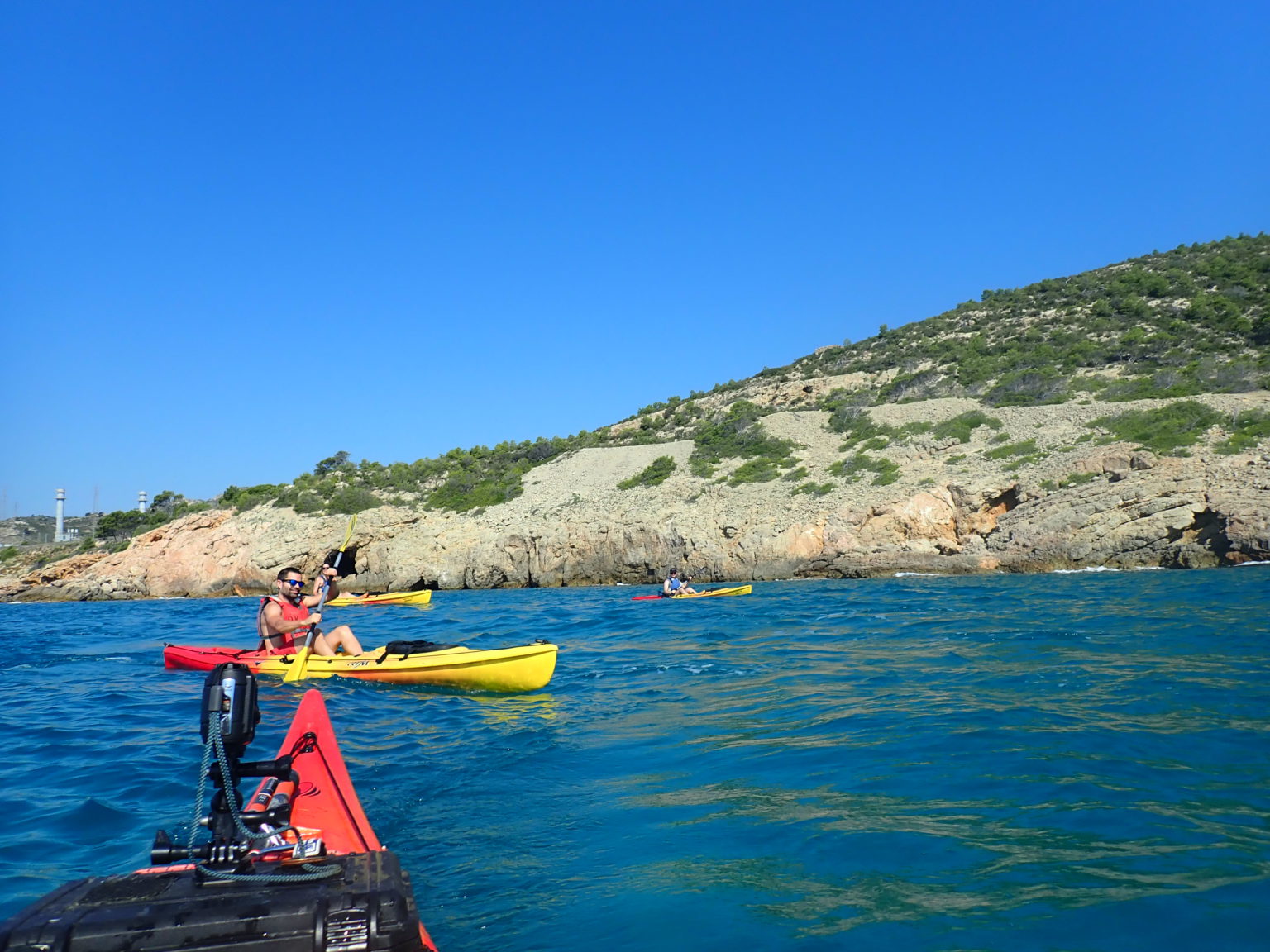 Un grupo de palistas en kayaks de colores navega por las aguas azules junto a una escarpada costa mediterránea.