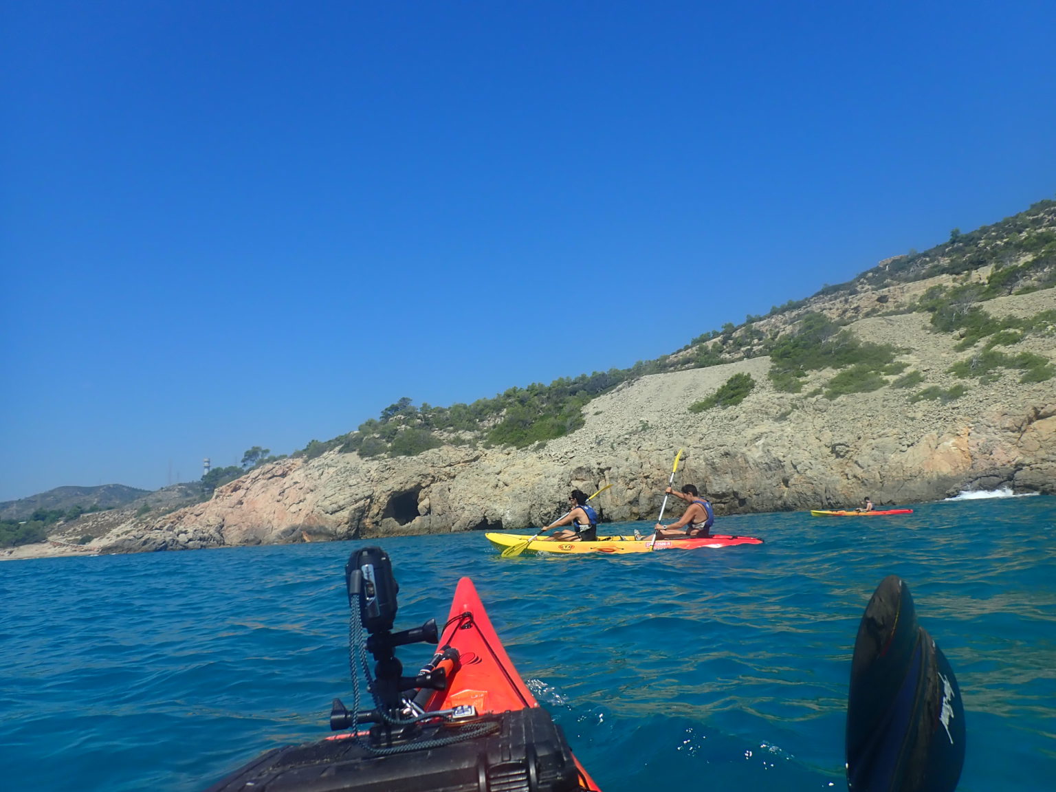 Vista desde un kayak de proa roja de un grupo de remeros explorando una costa rocosa bajo un cielo azul despejado.
