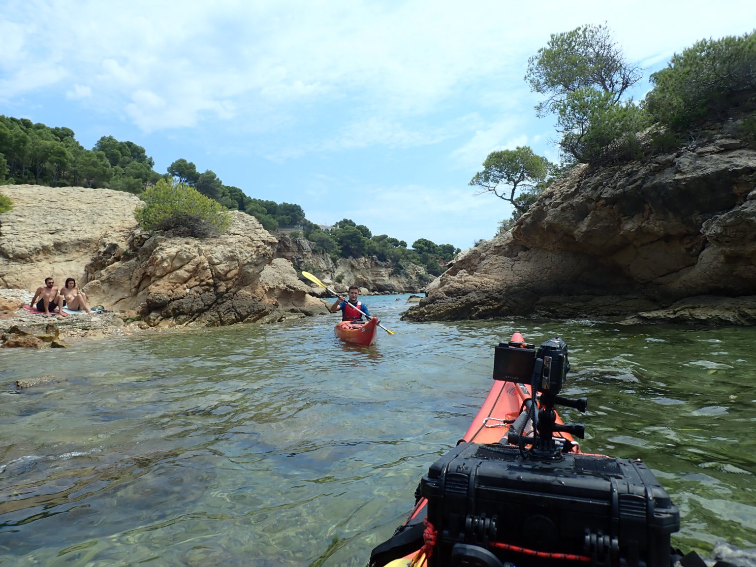 Vista desde un kayak de proa roja navegando en aguas transparentes hacia una persona en otro kayak, con formaciones rocosas y vegetación mediterránea a los lados.