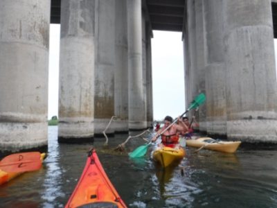 Un kayakista navegando entre los imponentes pilares de un puente, con restos de vegetación flotando en el agua.