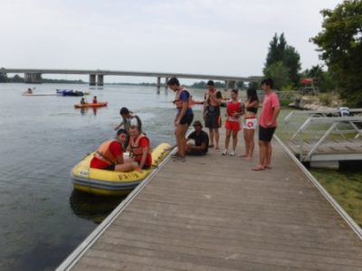 Un grupo de jóvenes se prepara para diversas actividades acuáticas en un muelle junto a un río, con botes inflables y kayaks visibles en el agua.