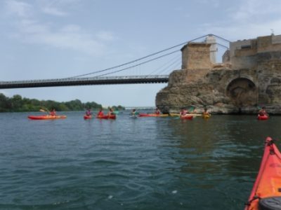 Un grupo de kayakistas navega por el río Ebro, pasando por debajo de un puente colgante y junto a una fortificación de piedra que se alza majestuosamente en la orilla derecha.