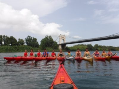 Un grupo diverso de palistas, incluyendo jóvenes y adultos, posan para una foto en sus kayaks individuales sobre el río Ebro, con un icónico puente colgante de fondo.