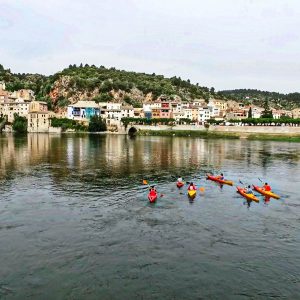 Kayaks navegando por el río Ebro con el pintoresco pueblo de Miravet y su castillo de fondo.
