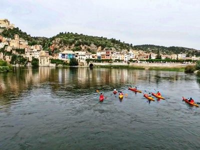 Kayaks navegando por el río Ebro con el pintoresco pueblo de Miravet y su castillo de fondo.