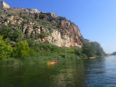 Un kayakista solitario navega por el Ebro con el imponente Castillo de Miravet dominando el horizonte rocoso, en una jornada de sol y naturaleza.