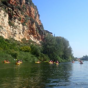 Grupo de kayaks navegando por las aguas del río Ebro, flanqueados por la imponente orilla rocosa y la exuberante vegetación ribereña.