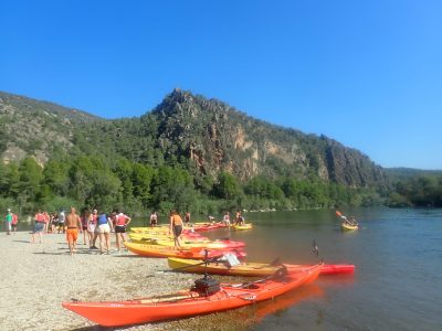 Un grupo de personas se prepara para una emocionante jornada de kayak en un amplio río, con una flota de embarcaciones coloridas en la orilla y majestuosas montañas como telón de fondo.